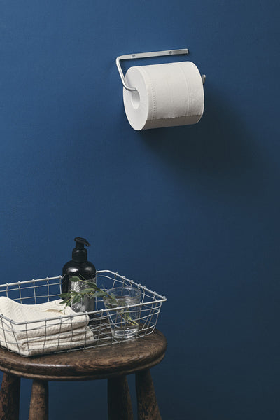 White toilet paper roll on a holder against a blue wall with a wooden stool and basket containing towels and a bottle.