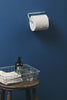 White toilet paper roll on a holder against a blue wall with a wooden stool and basket containing towels and a bottle.